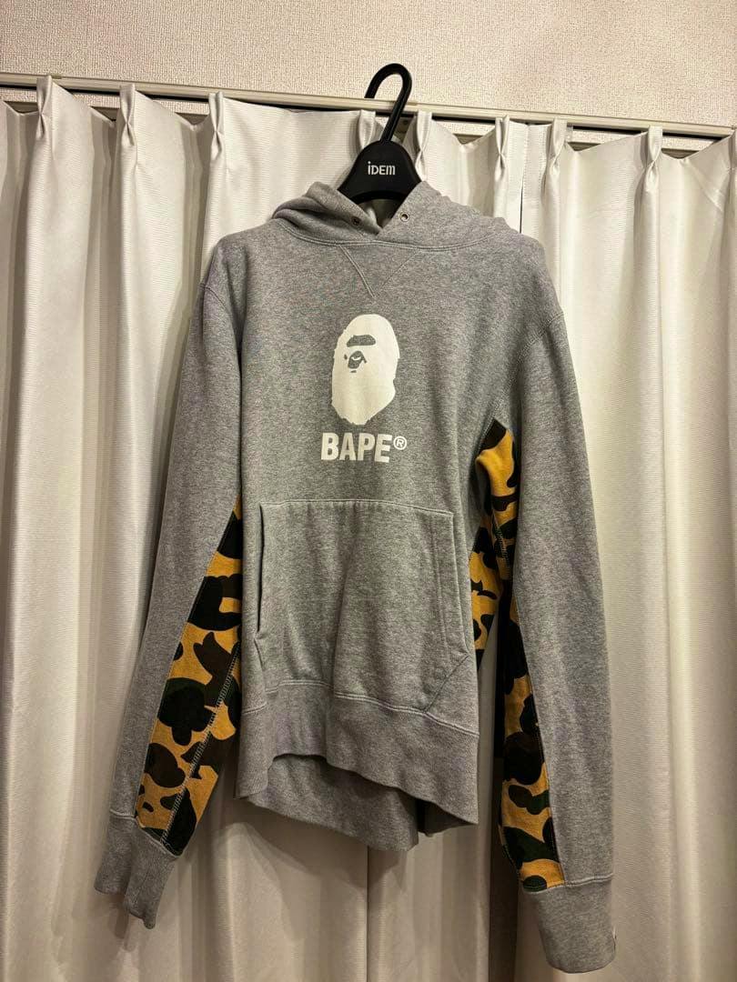 Gray A Bathing Ape hoodie with a white ape head logo and "BAPE" text on the chest and yellow-green camouflage panels on the sleeves, hanging on a hanger.