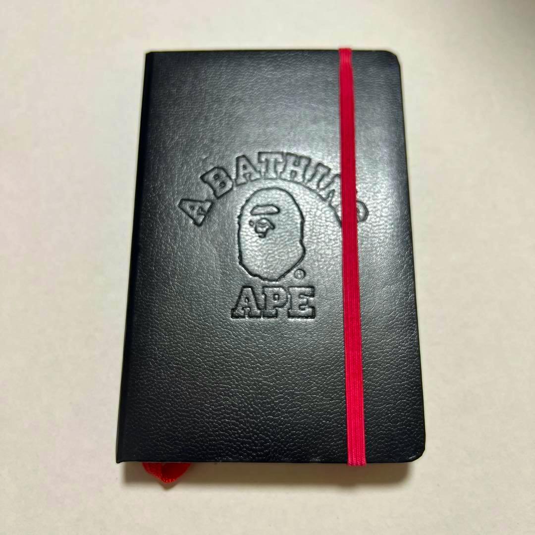 Black A BATHING APE notebook with embossed logo on the cover and a red elastic band, shown on a light background.