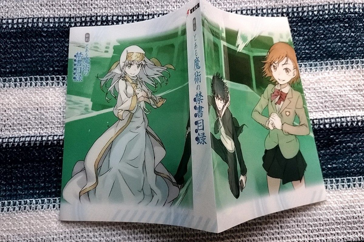 Clear illustrated book cover for "新約 とある魔術の禁書目録" showing three characters on a green background, photographed laid flat.