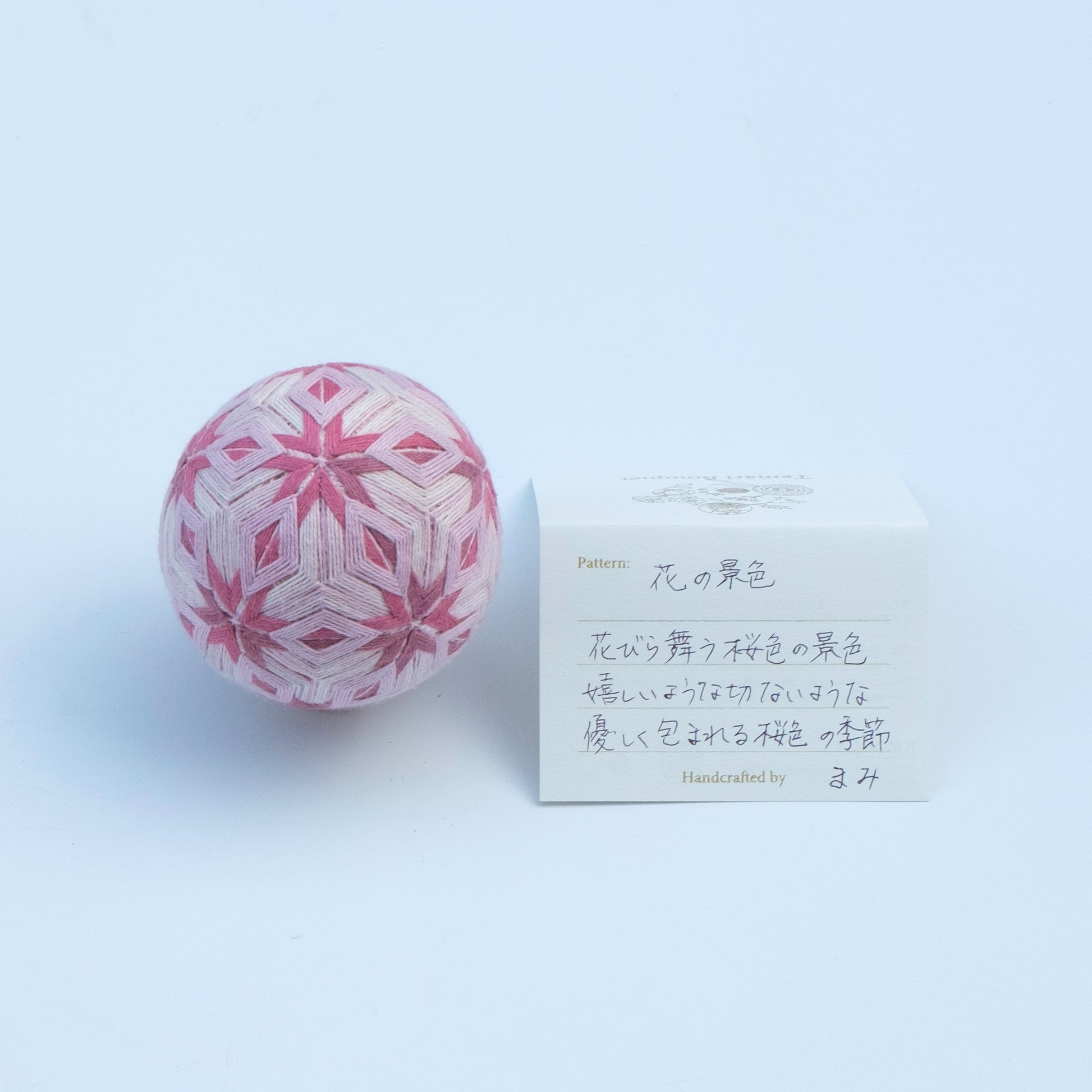 A pink and white embroidered temari ornament shown next to a small handwritten card.