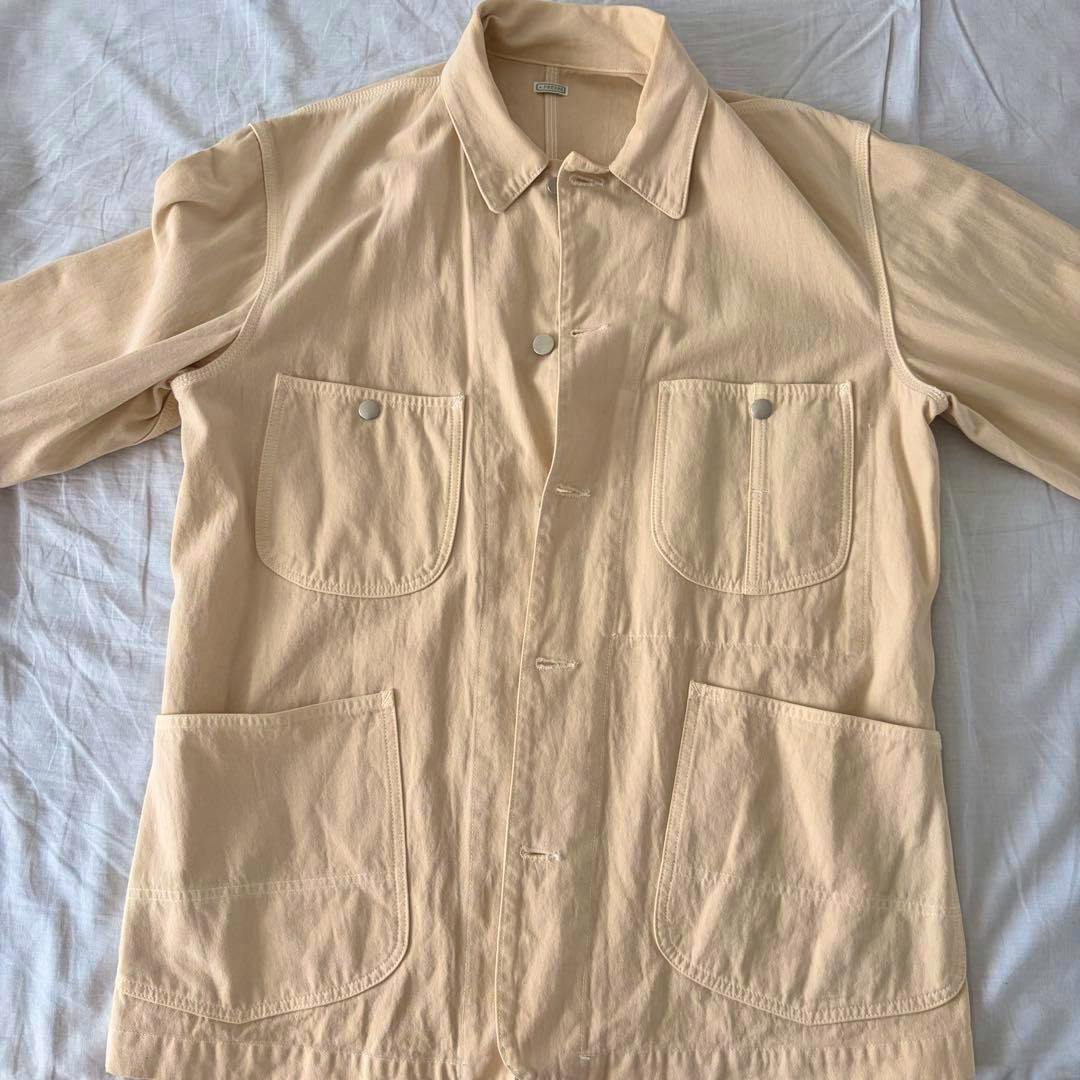 Beige cotton coverall jacket laid flat showing the front with four patch pockets and button closures.