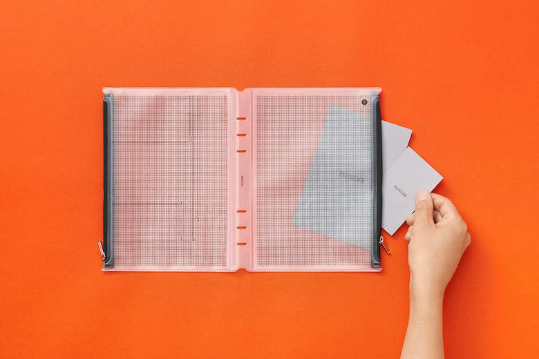 Opened A5 zipper case laid flat on an orange background with a hand inserting cards into the right pocket.