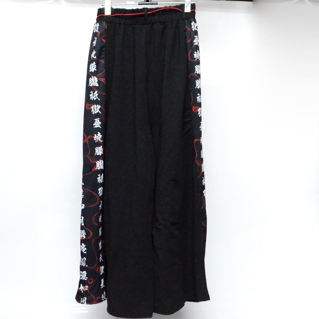 Front view of black hakama-style wide polyester pants with white and red kanji printed side panels and elastic waistband.