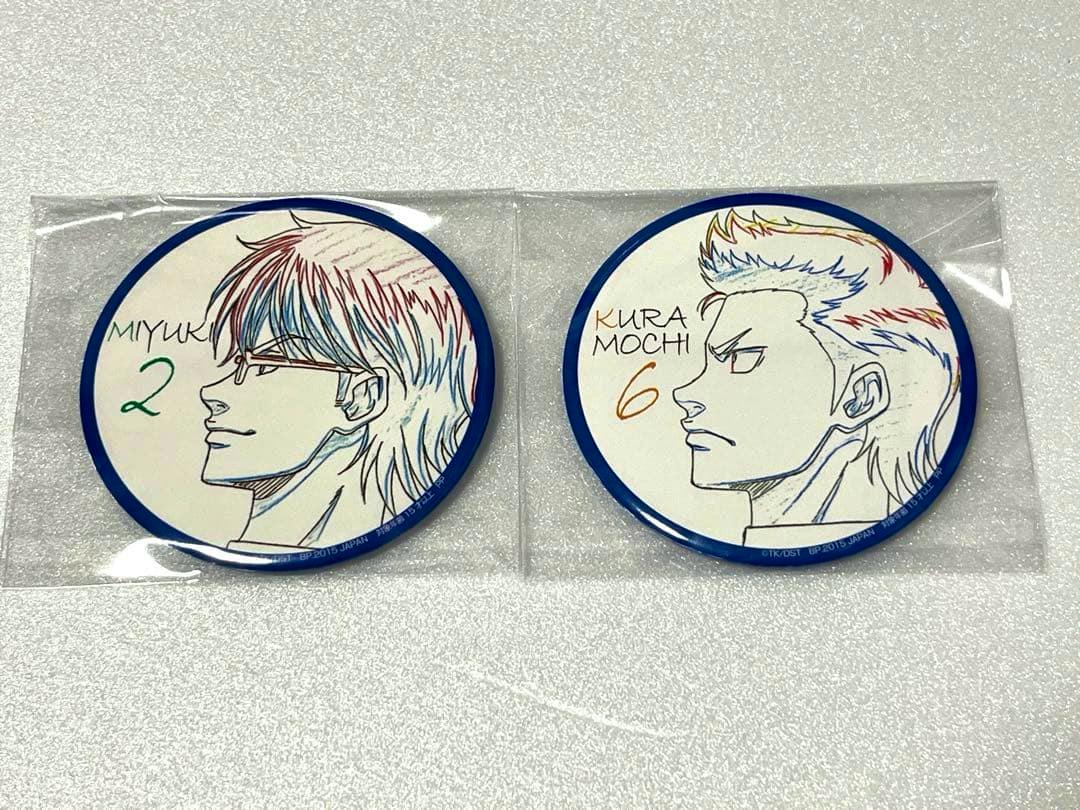 Two round can badges showing side-profile sketches of Miyuki and Kuramochi in individual plastic sleeves on a light surface.