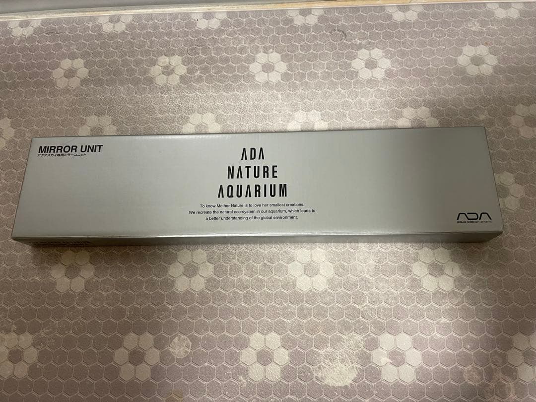 Silver rectangular box labeled "ADA NATURE AQUARIUM" mirror unit photographed on a patterned surface.