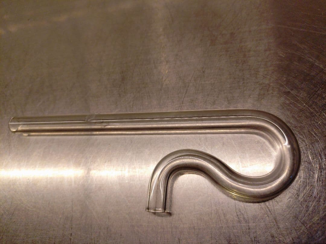 Glass aquarium intake lily pipe (ADA リリィパイプ ミニ p-1, ∅10) lying on a metal surface, showing the S-shaped curved outlet and straight inlet tube.