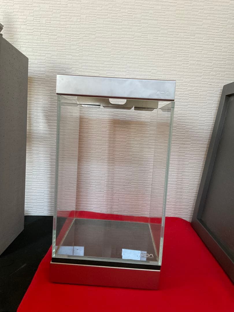 Clear ADA Mini NA glass aquarium with metal lid and base on a red surface.