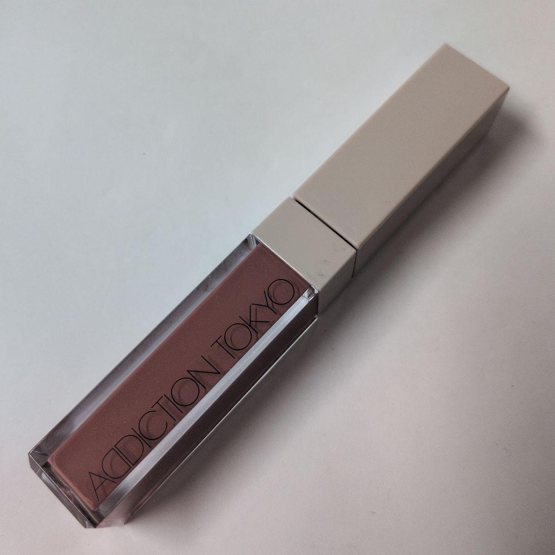 A tube of ADDICTION TOKYO liquid lip gloss in a clear rectangular container with a pale pink product and a white square cap on a white background.