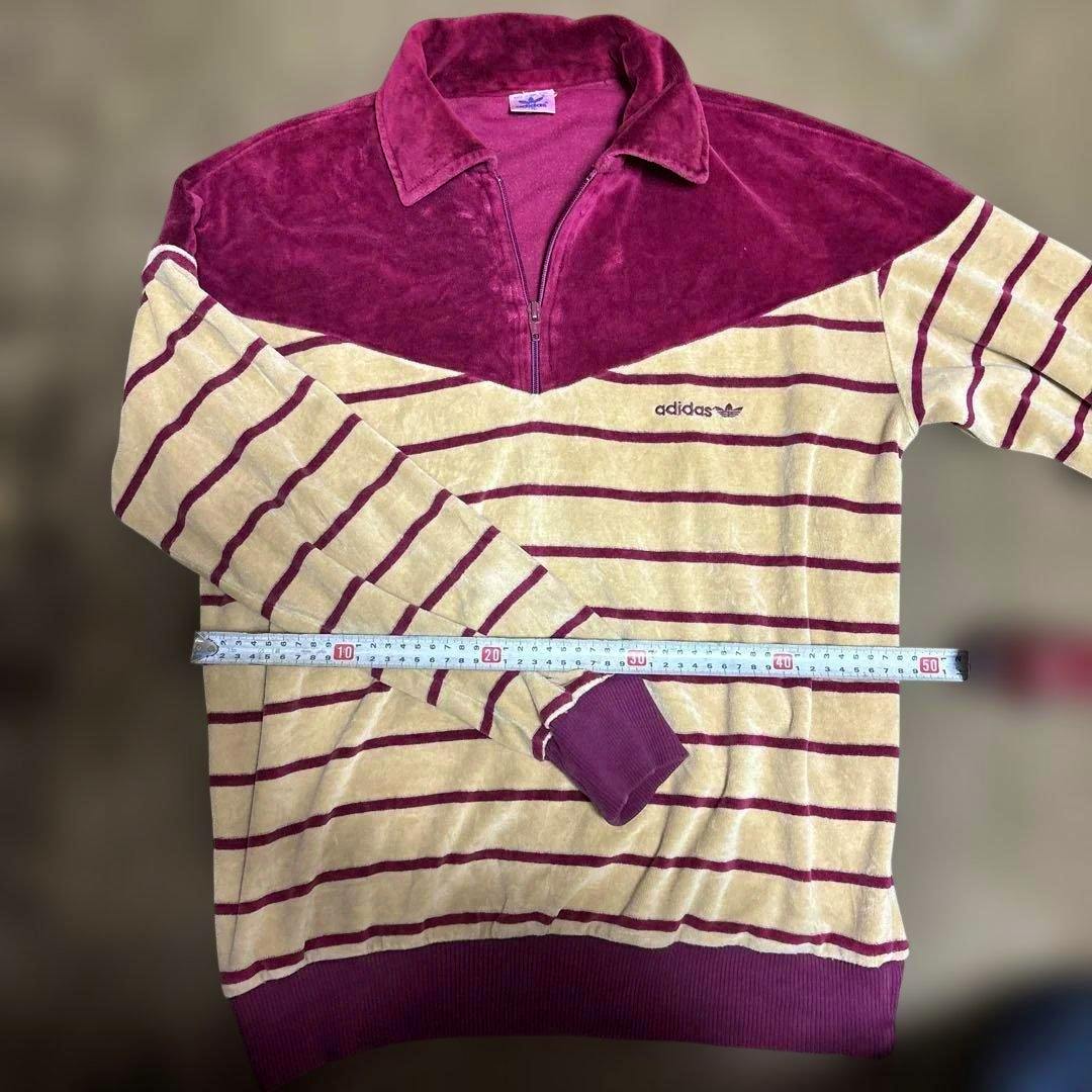 Flat-lay photo of an adidas striped zip-up trainer with a maroon collar and beige-and-maroon horizontal stripes, with a measuring tape across the chest.