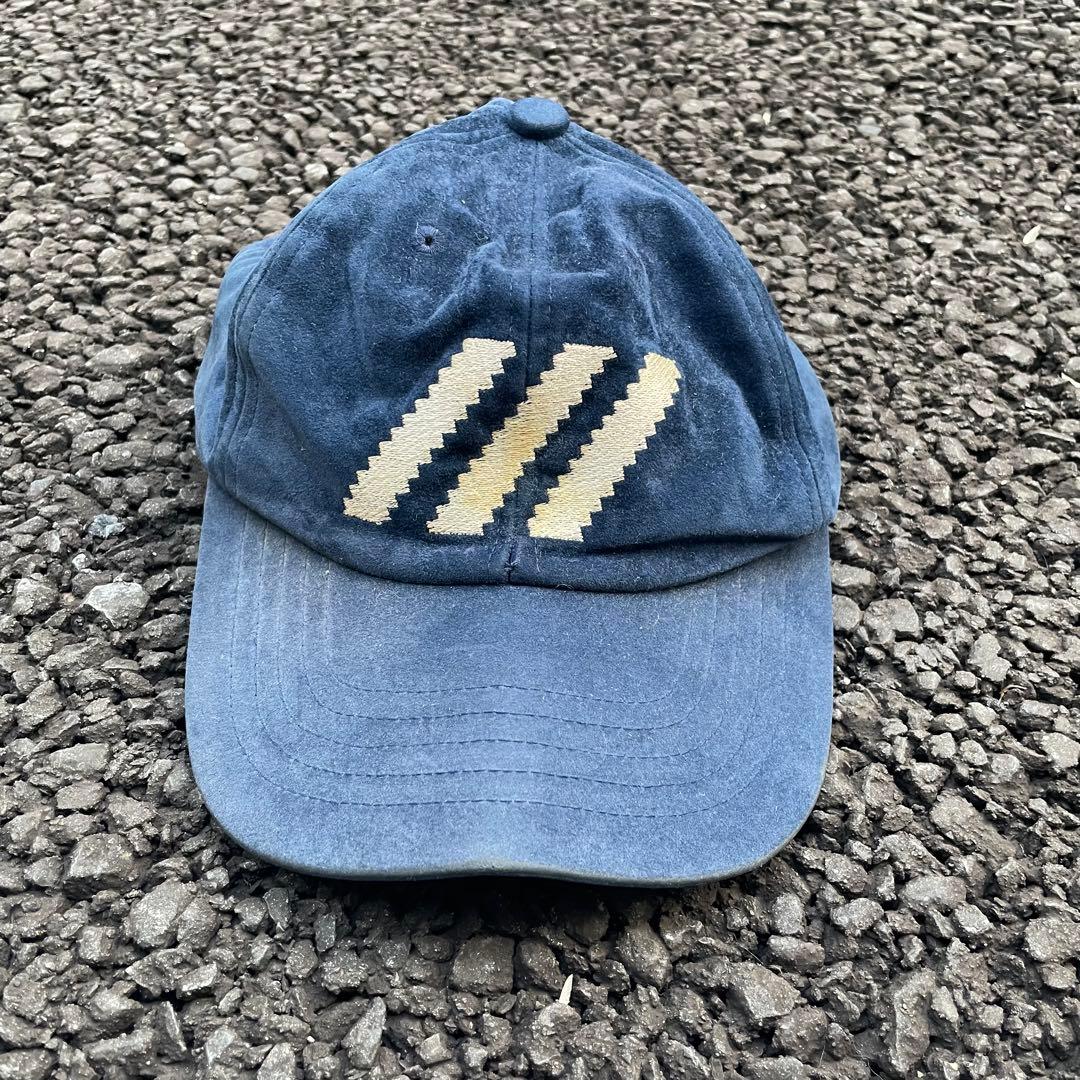 Front view of a navy adidas polyester cap with three diagonal white stripes, photographed on asphalt.