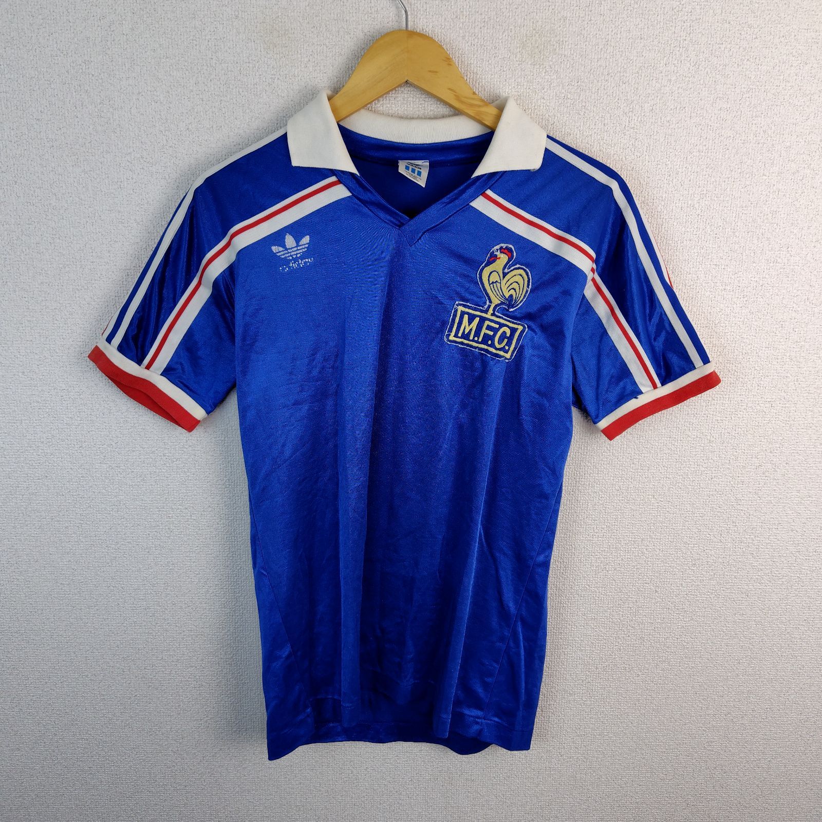 Blue adidas M.F.C. short-sleeve soccer game shirt on a hanger against a wall.