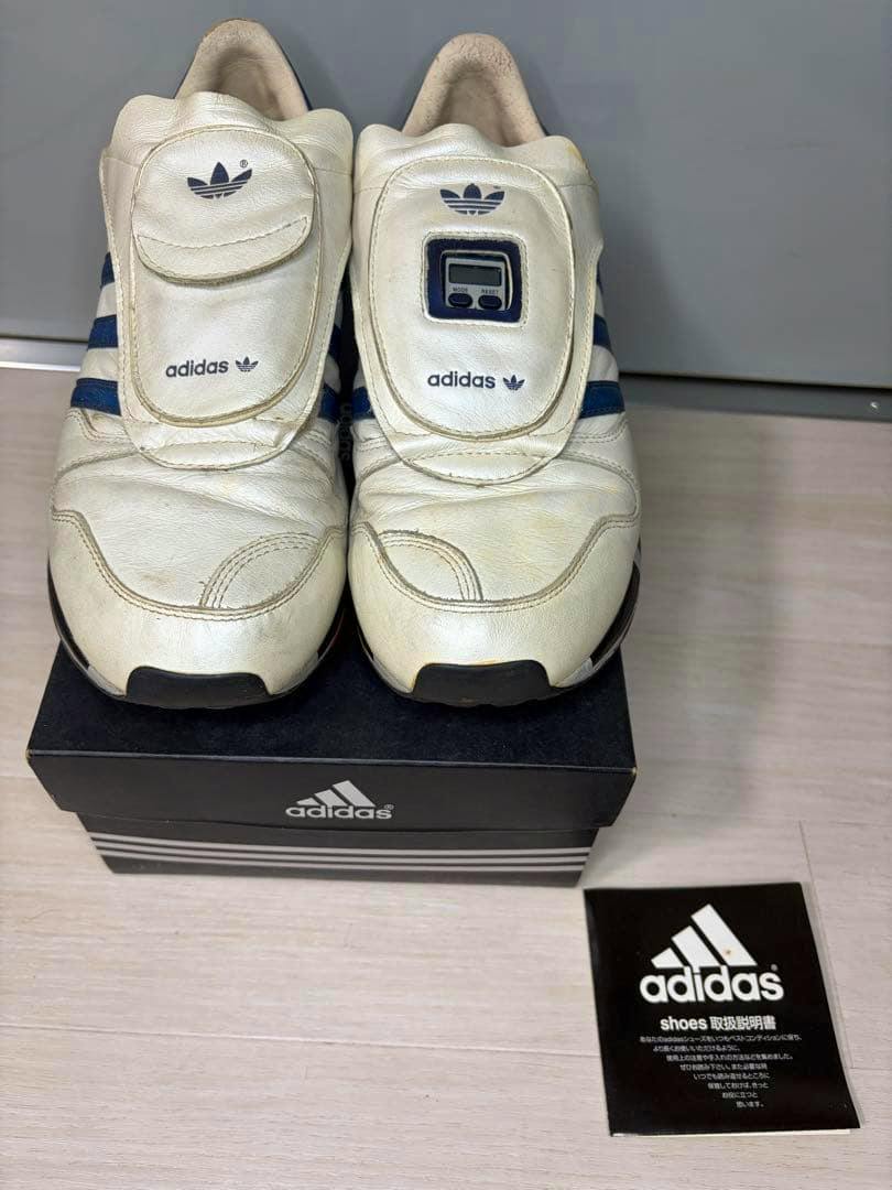 A pair of white adidas MICROPACER2 sneakers with blue stripes and a small digital display on the tongue, placed on an adidas shoebox with an adidas booklet beside them.