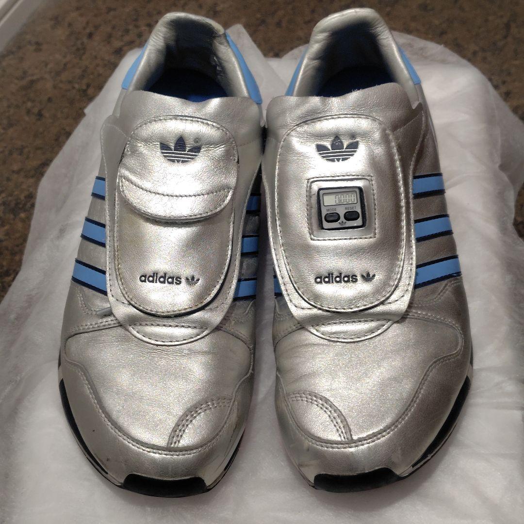 Top-down view of a pair of silver adidas MICROPACER leather shoes with a built-in digital display on the tongue.