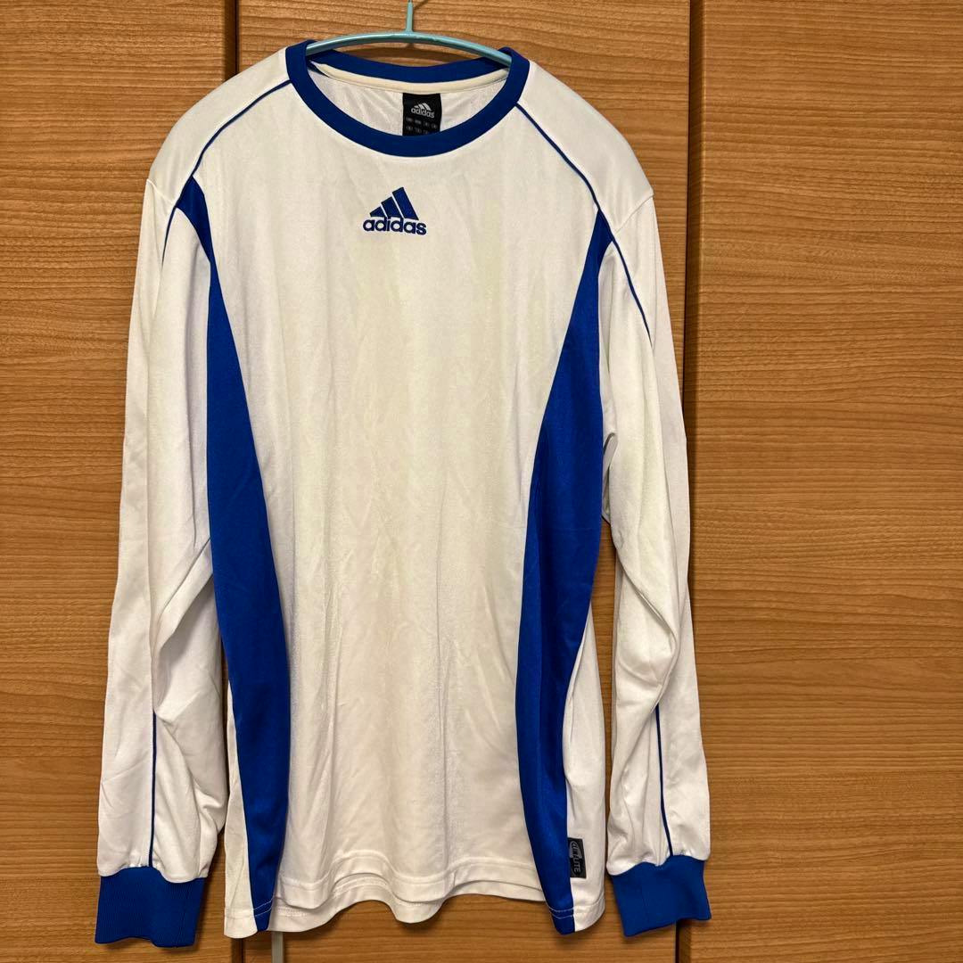 White and blue adidas long-sleeve soccer jersey hanging on a hanger against a wooden background.