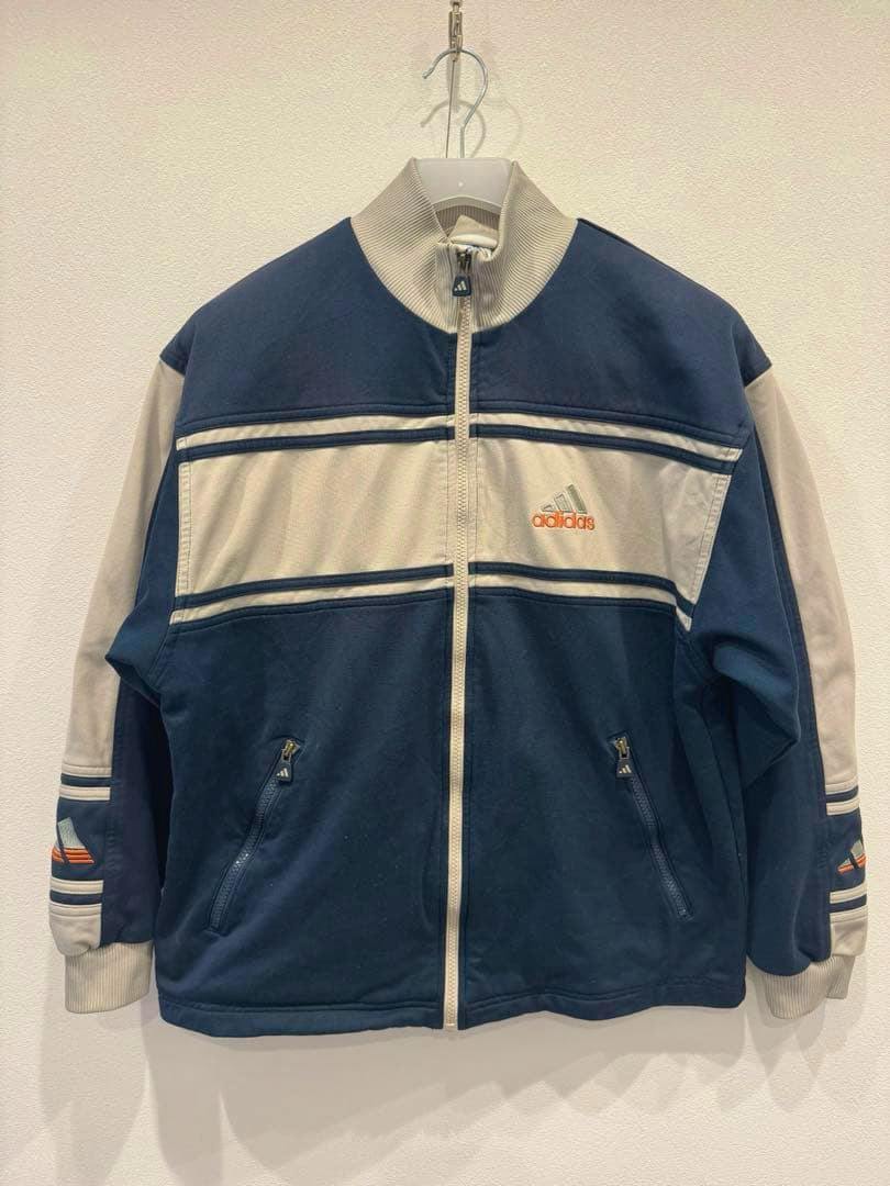 Front view of the used Adidas track jacket in navy and beige with a center zipper and embroidered Adidas logo hanging on a wall.
