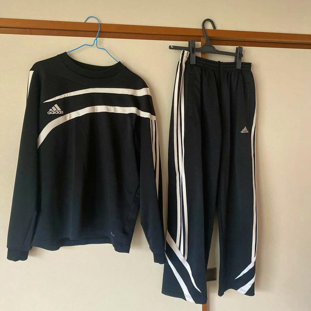 Black adidas training top and matching pants with white stripes hanging on hangers against a wall.