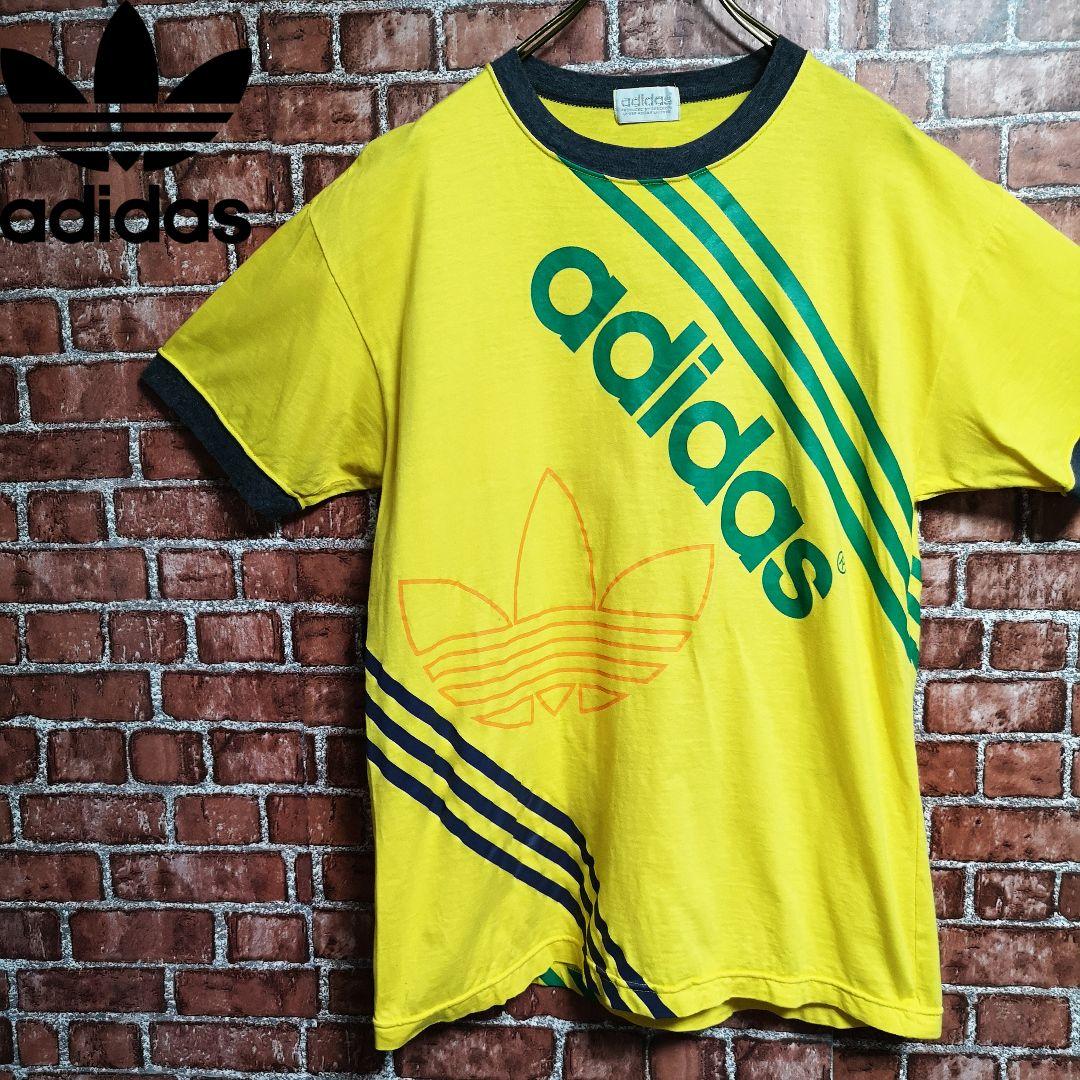 Yellow men's polyester Adidas short-sleeve T-shirt with diagonal green “adidas” text and trefoil design, hanging on a hanger against a brick wall.