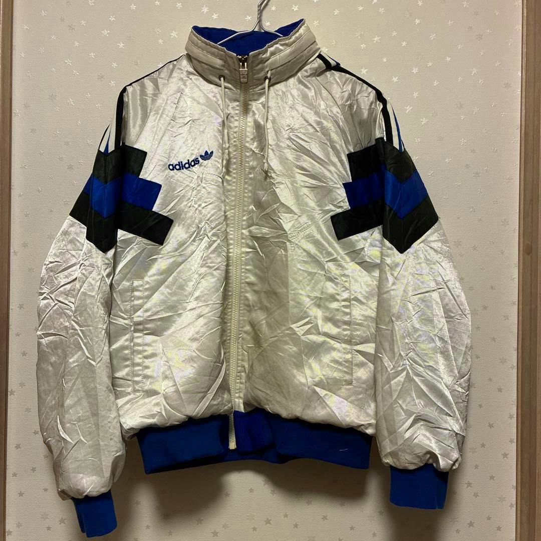 Front view of a white vintage adidas nylon men's jacket with blue and black chevron sleeve details hanging on a hanger.