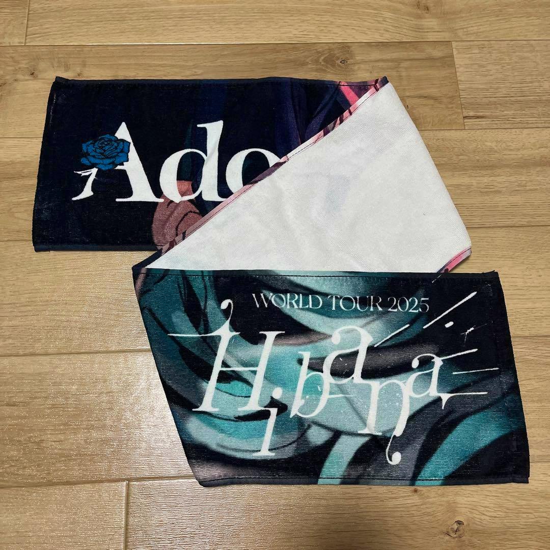 Photo of the Ado "Hibana" muffler towel folded on a wooden floor showing the printed Ado and Hibana designs.