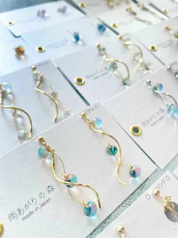 Pair of curved gold-tone earrings with blue-green glass beads displayed on a white product card labeled '雨あがりの森'.