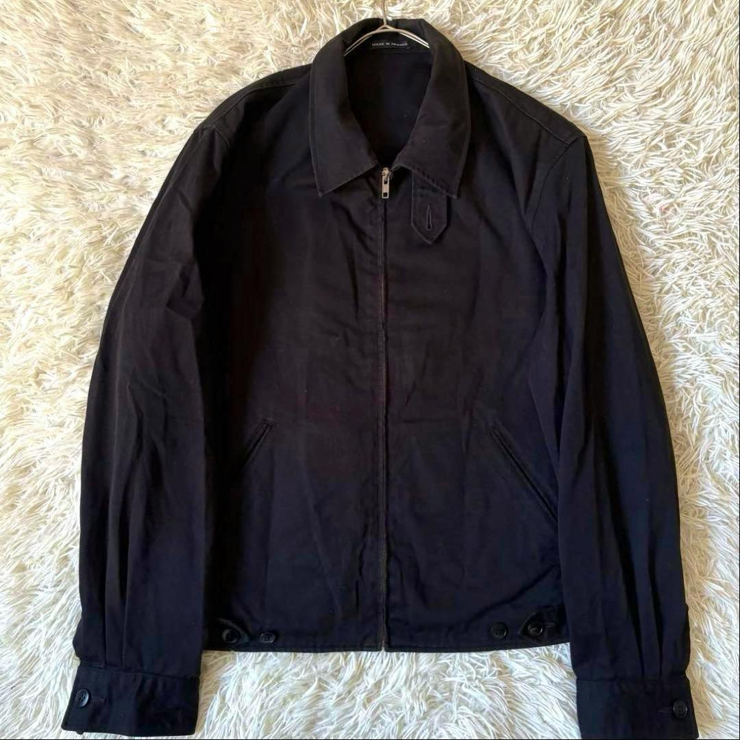Front view of a black agnes b. homme swing-top men's cotton jacket laid flat on a light shag rug, showing the zipper and collar.