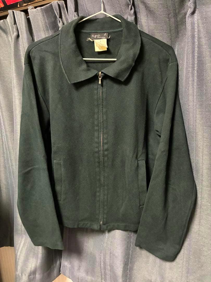 Front view of an agnes b. HOMME cotton blouson hanging on a hanger against a curtain.