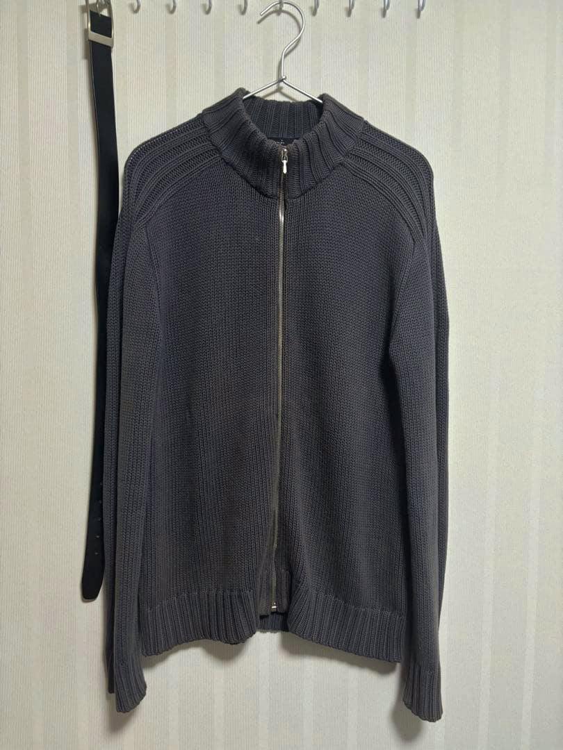 Front view of an agnes b. men's zip-up driver's knit sweater hanging on a hanger against a wall.