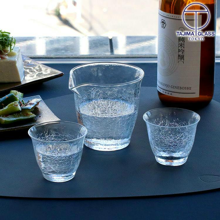 Cracked-texture glass cold-sake set showing one katakuchi pourer and two guinomi cups on a dark placemat with a bottle and dishes in the background.