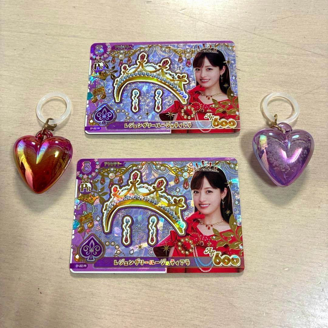 Two holographic Aikatsu collectible cards showing a woman with a crown motif, alongside two heart-shaped keychain charms on a light wood surface.