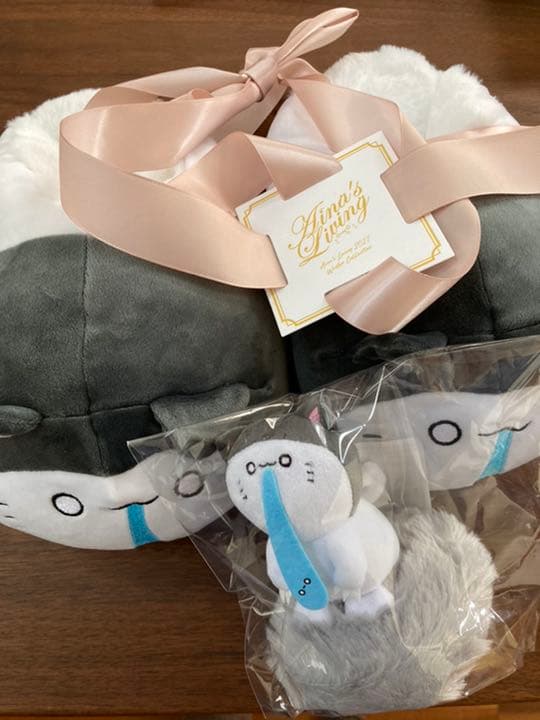 Grey plush room slipper with a pink ribbon Aina's Living tag and a small packaged plush accessory on a wooden surface.