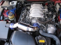 Metallic air control chamber with blue silicone couplers installed in a car engine bay.