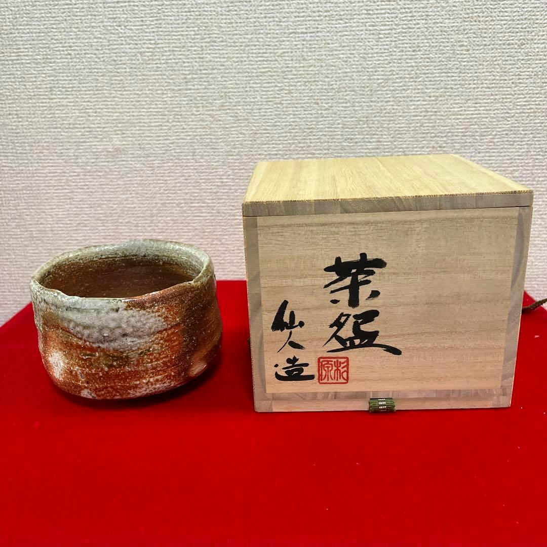 A ceramic matcha tea bowl displayed next to a wooden storage box on a red cloth.