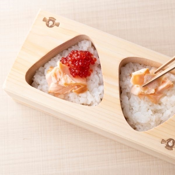 Wooden onigiri mold with two triangular cavities holding rice topped with salmon and salmon roe.