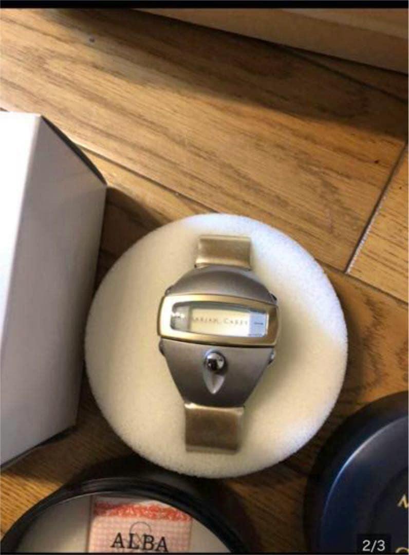 Close-up of a gold-tone ALBA Mariah Carey watch on a round foam display with its box and warranty case partially visible on a wooden floor.