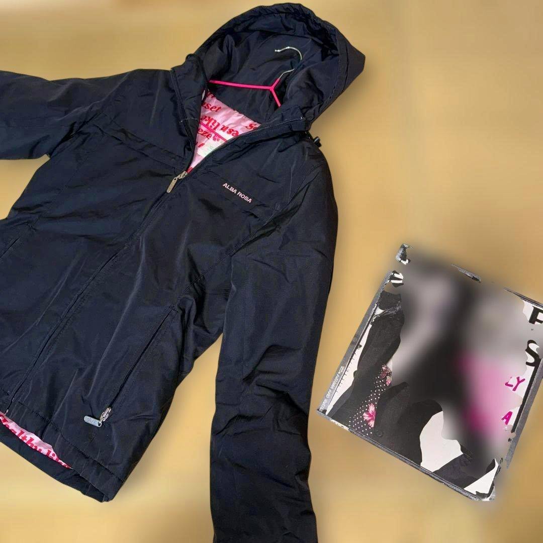 Black ALBA ROSA hooded women's jacket laid flat showing the front zipper and hood with a blurred magazine beside it.
