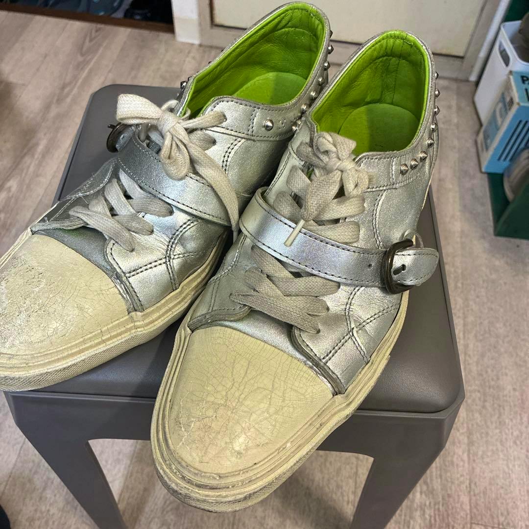 アルフレッドバニスタースニーカー: pair of metallic silver sneakers with bright green inner lining, studded heel, buckled strap, and worn beige toe caps placed on a small stool.