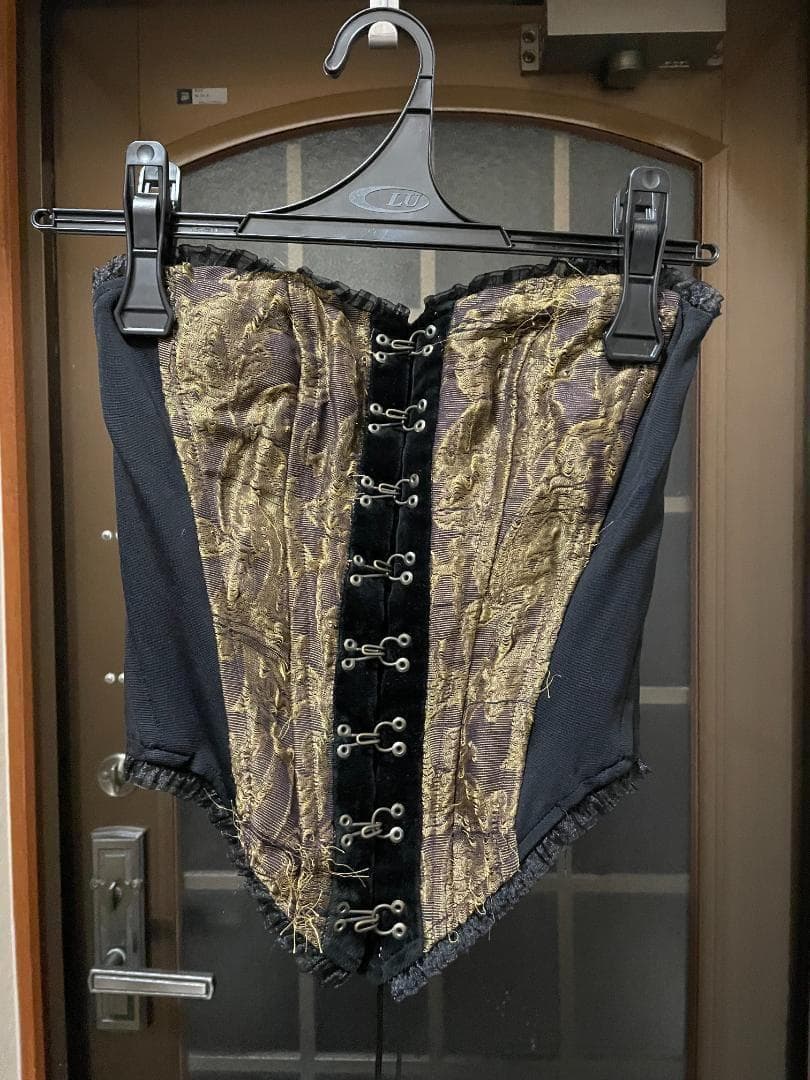 Front view of a black and gold gobelin-weave corset hanging on a hanger.