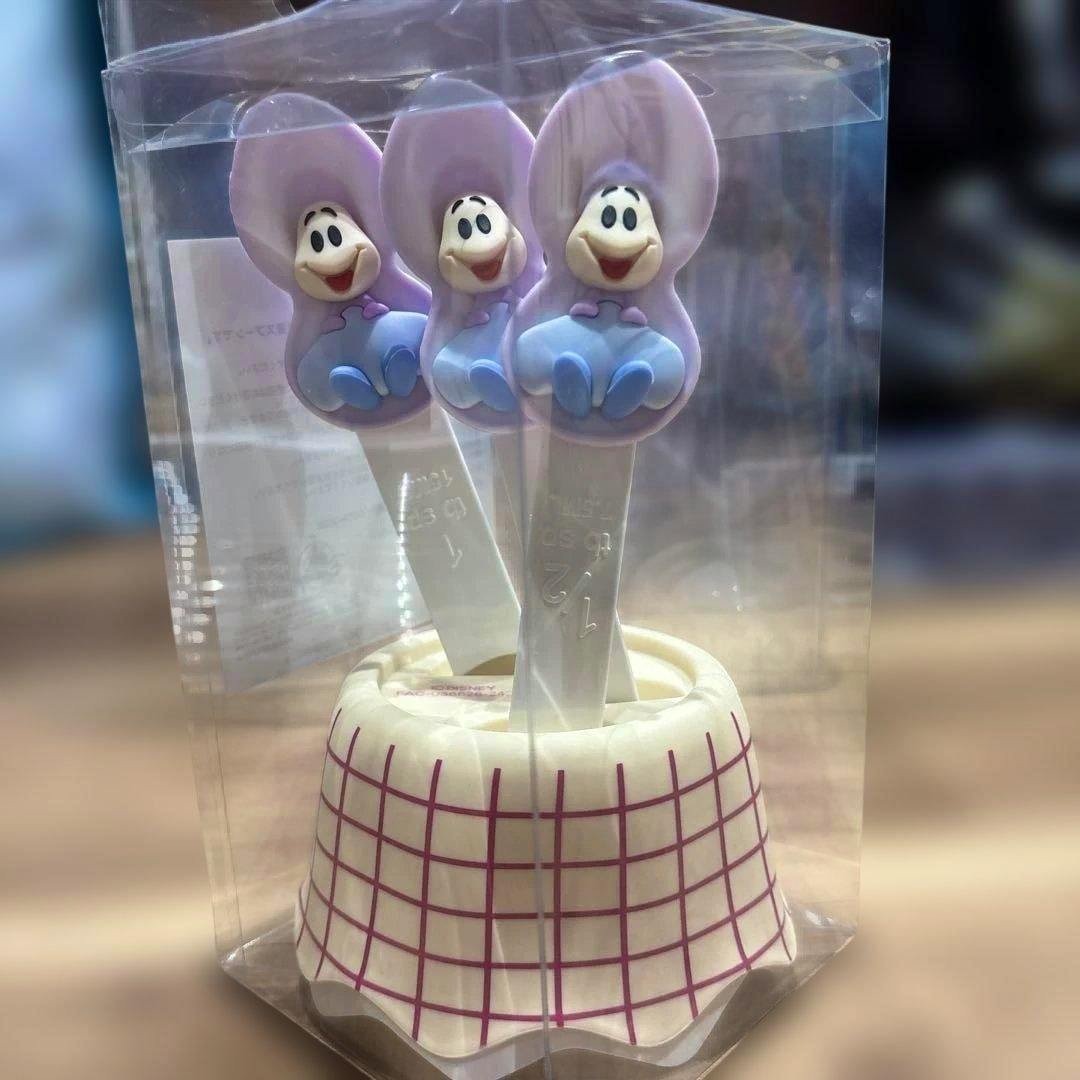 Three plastic measuring spoons with purple character tops displayed upright in clear retail packaging with a white checkered base.