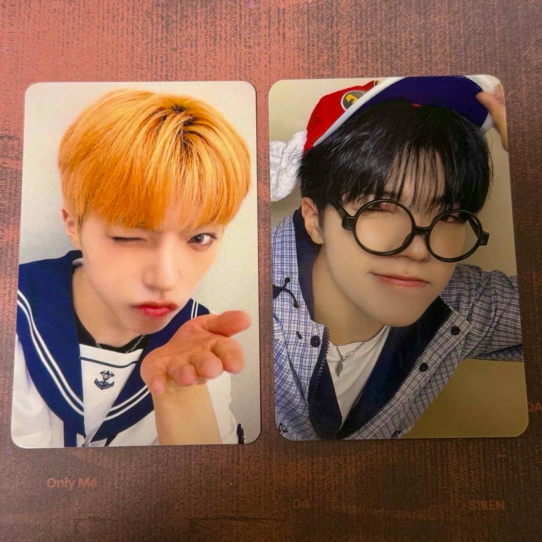 Two photocards side-by-side: left shows a person with orange hair winking and blowing a kiss in a sailor-style outfit; right shows a person with black hair wearing round glasses and a cap.
