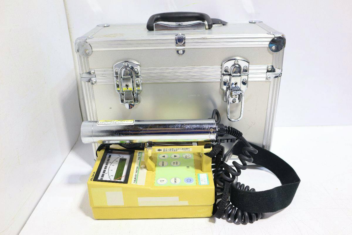 Yellow Aloka TCS-172 survey meter with attached probe and coiled cable in front of a silver carrying case.