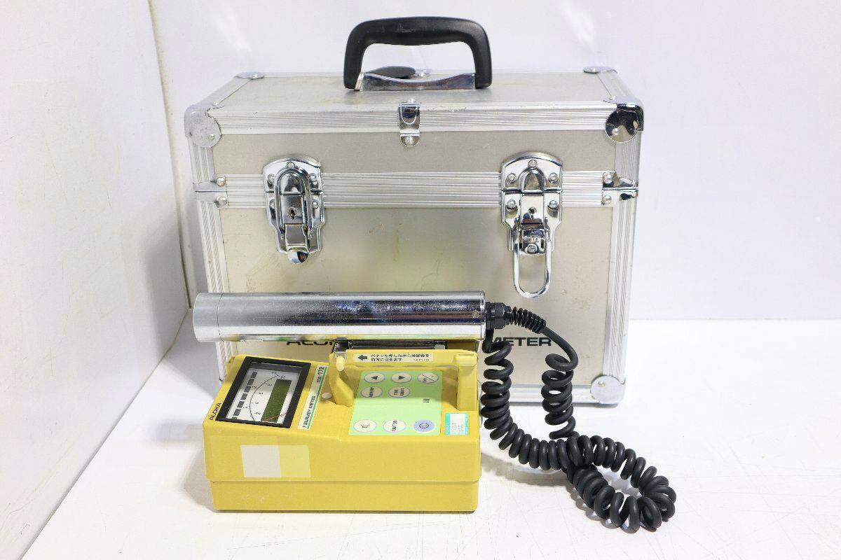 Yellow ALOKA TCS-172 survey meter with a cylindrical probe and coiled cable placed in front of a silver carrying case.