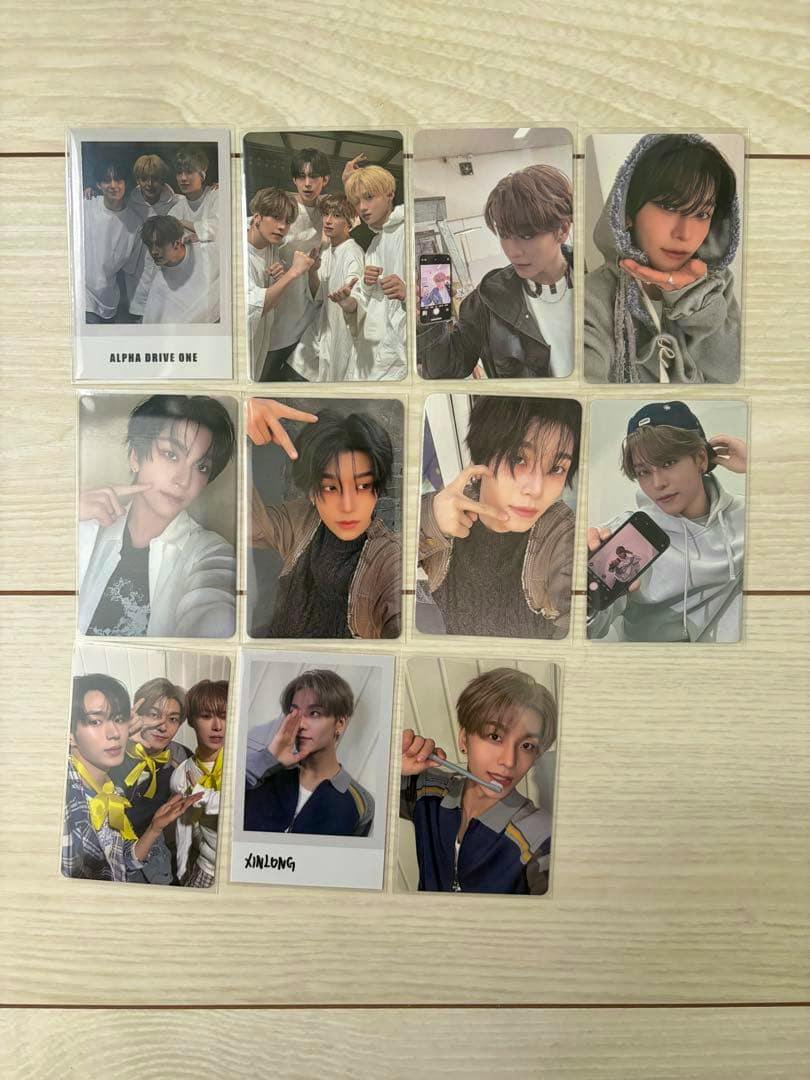 Several ALPHA DRIVE ONE XINLONG photocard images arranged in a grid on a light wood surface.