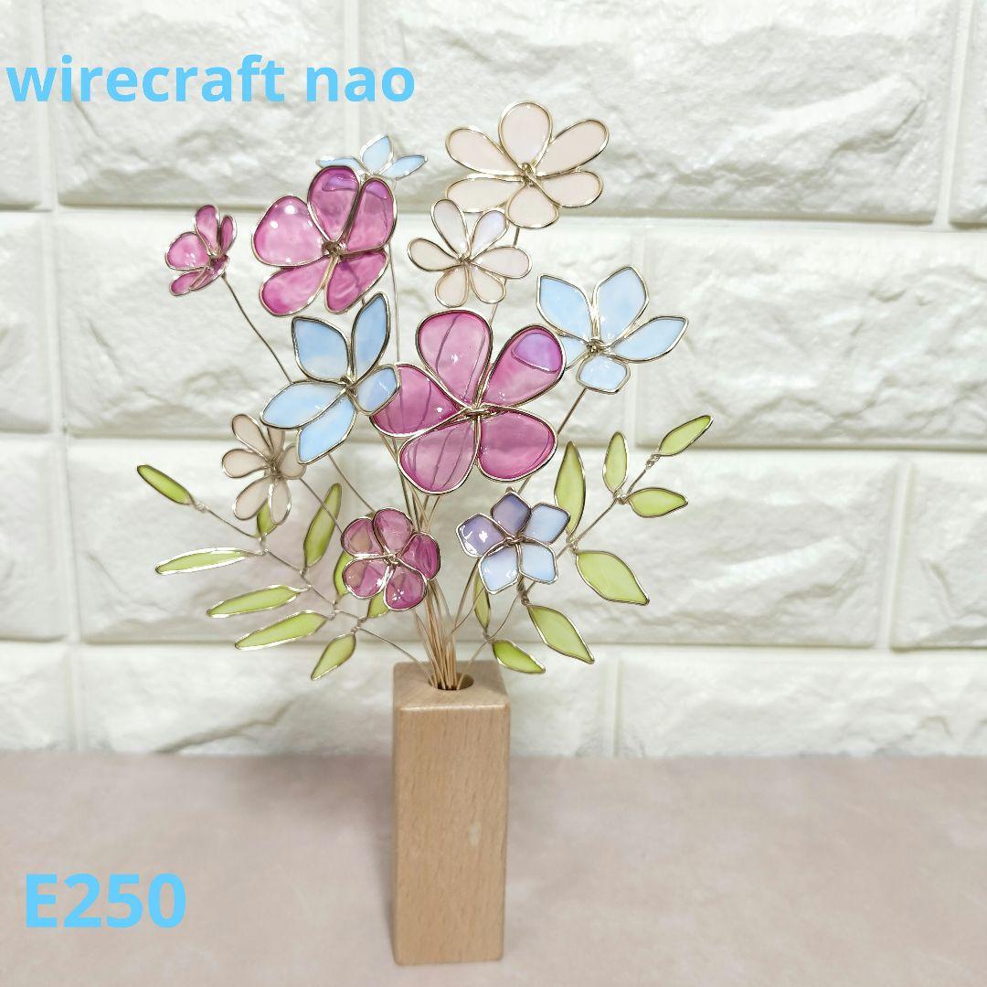 Wire and aluminum flower arrangement with pink, blue, and cream petals displayed in a small wooden block vase against a white tiled background.