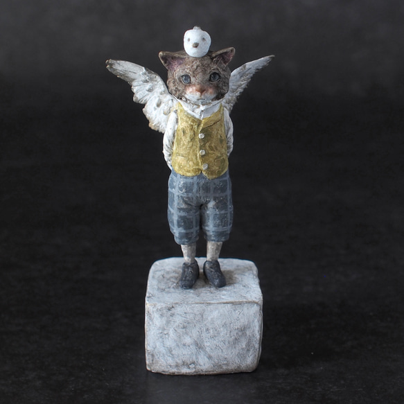 Front view of a ceramic figure of a winged cat wearing a yellow vest and standing on a cube base.