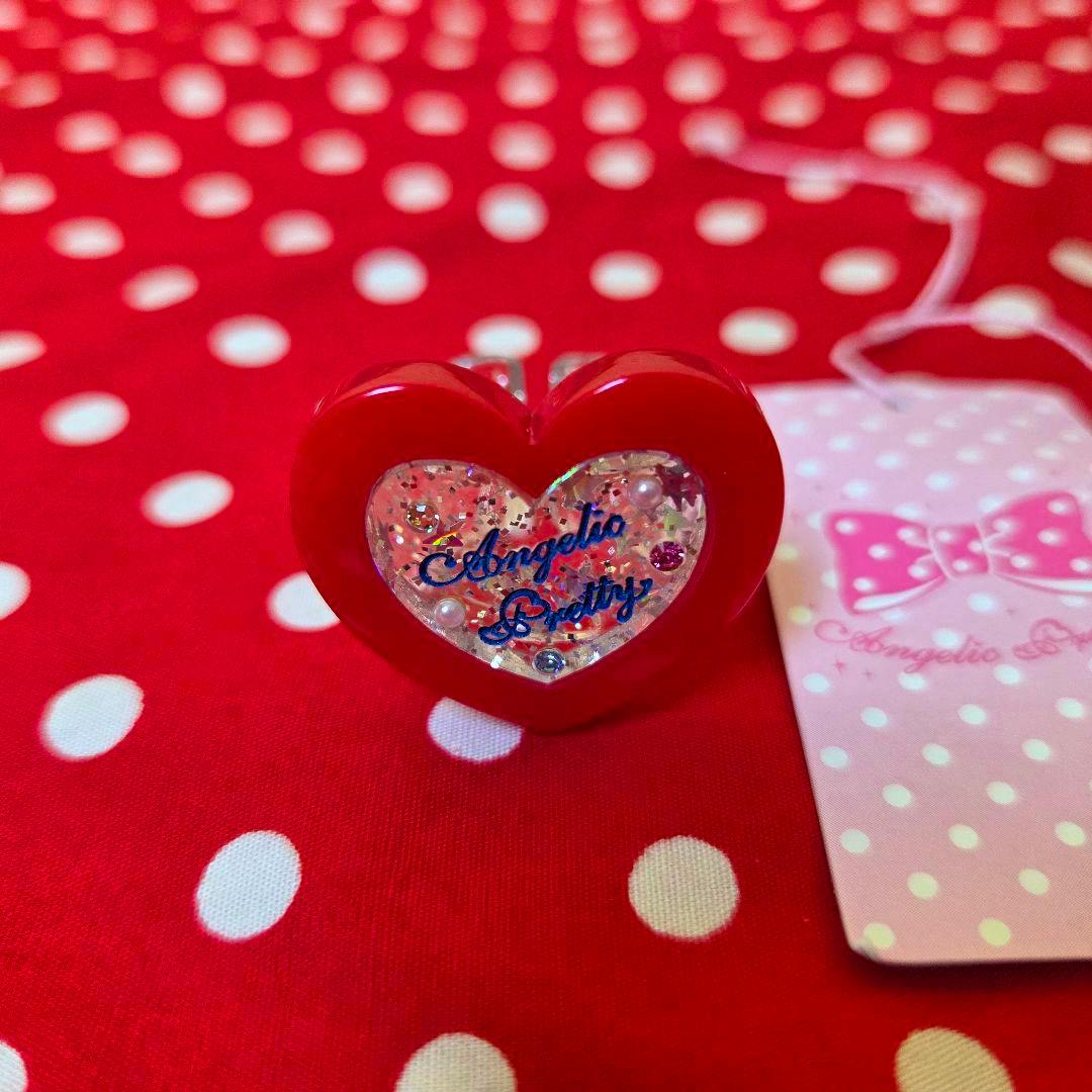 Red heart-shaped plastic ring with a glittery clear center reading "Angelic Pretty" on a red polka-dot background.
