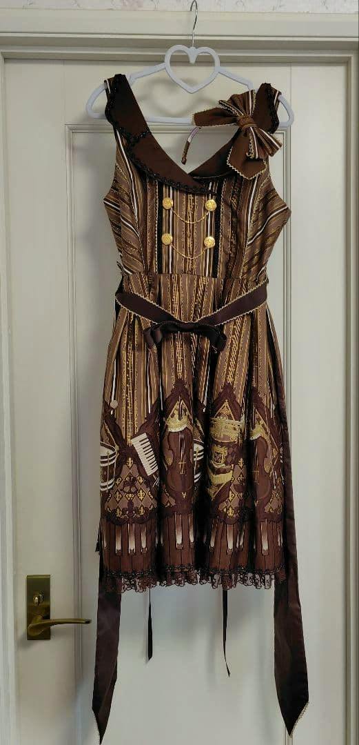 Brown Angelicpretty collar JSK jumper skirt with striped and musical-print pattern, gold decorative buttons, bow, and long ribbons hanging on a heart-shaped hanger against a door.