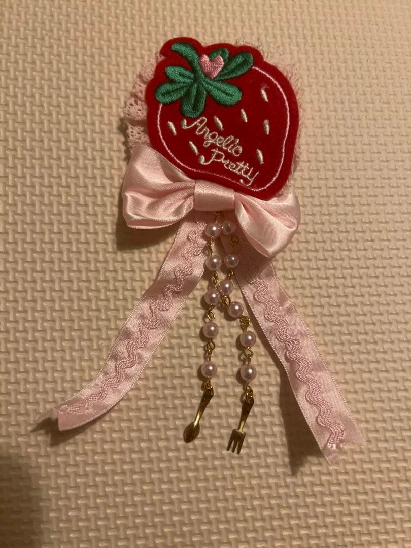Pink Angelic Pretty strawberry patch brooch with a bow, lace ribbons, and dangling beaded chains with small utensil charms on a textured background.