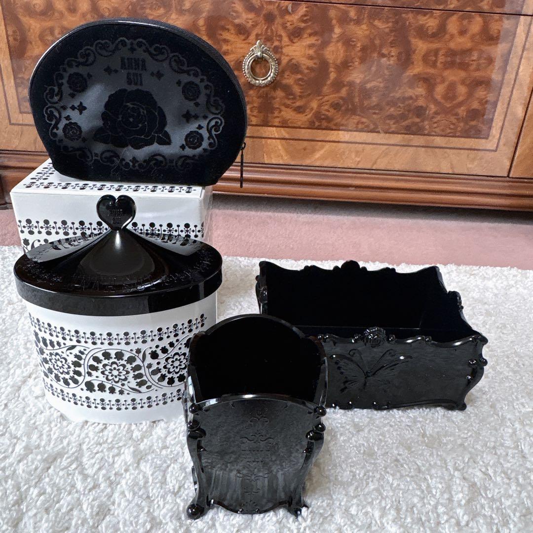 Anna Sui beauty box pouch set pieces — a white-and-black round box with lid and two black ornate plastic containers arranged on a carpet.