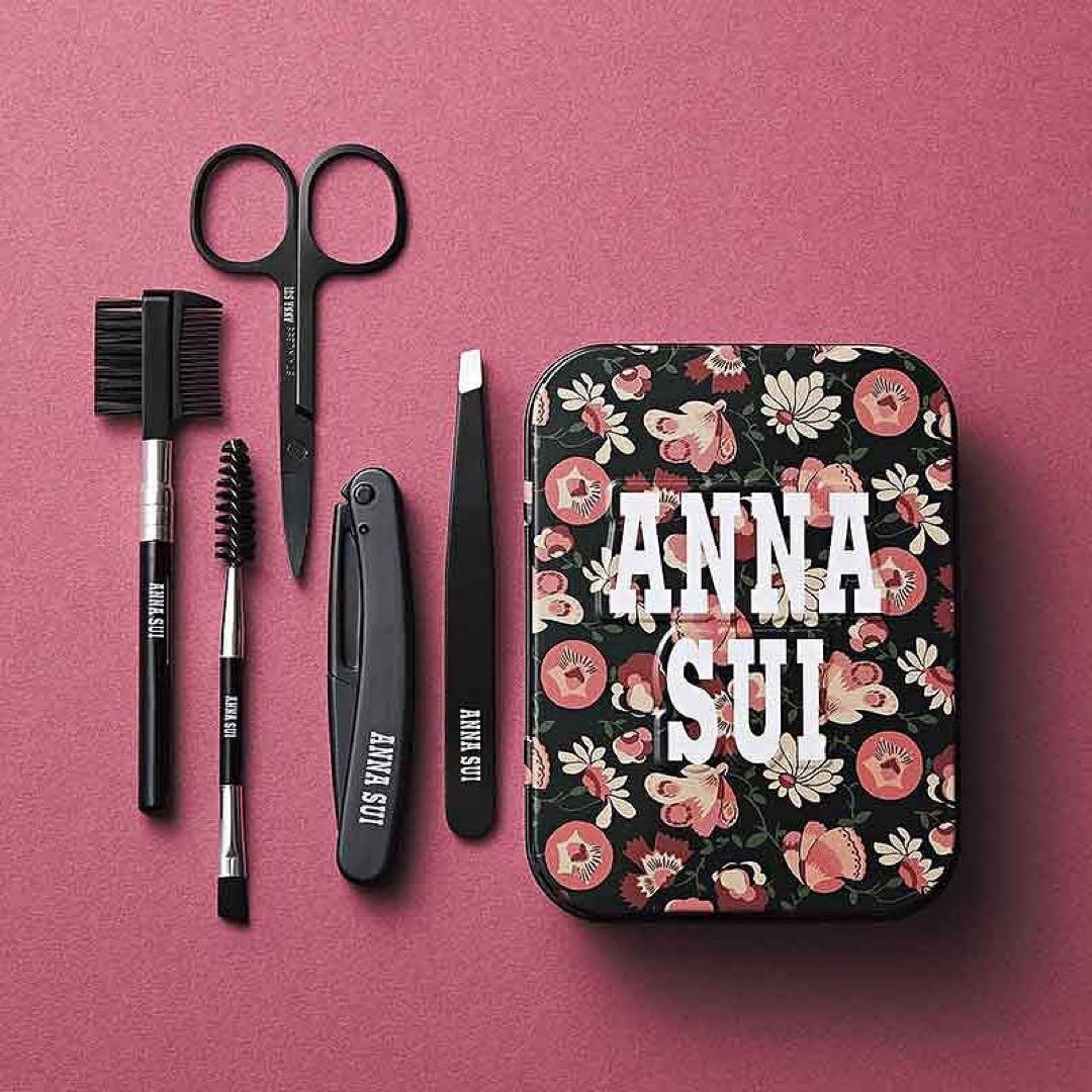 Five-piece eyebrow grooming set displayed next to a floral ANNA SUI tin case, showing scissors, tweezers, a spoolie, and two small brow brushes.