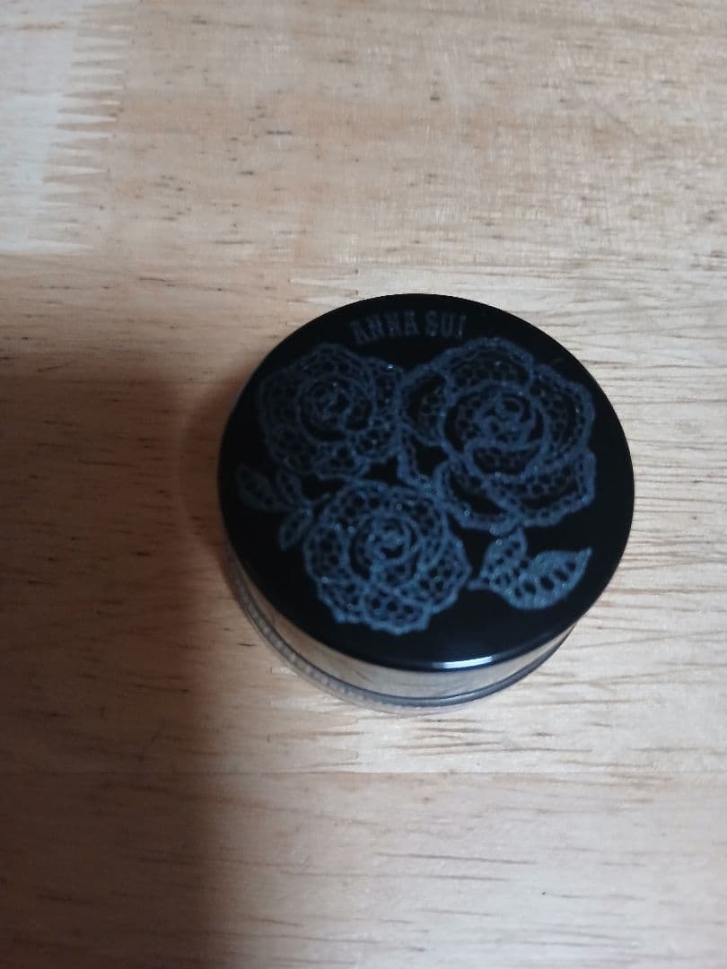 Closed ANNA SUI mousse eyeshadow pot with a black floral-patterned lid on a wooden surface.